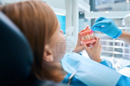 Junge Patientin untersucht Kieferattrappe mit Zahnspange, Zahnarzt bringt Kind Mundhygiene bei