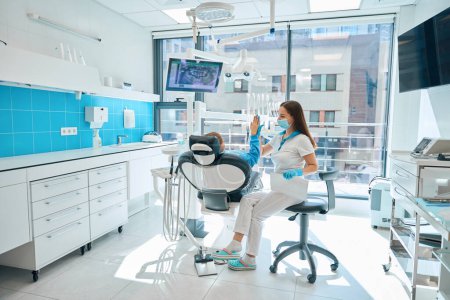 Photo for Dentist in a protective mask greets a girl at a consultation, a child in a blue sweatshirt - Royalty Free Image