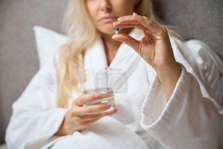Photo for Cropped photo of woman in bathrobe seated in bed holding glass of water and medicine capsule in hands - Royalty Free Image