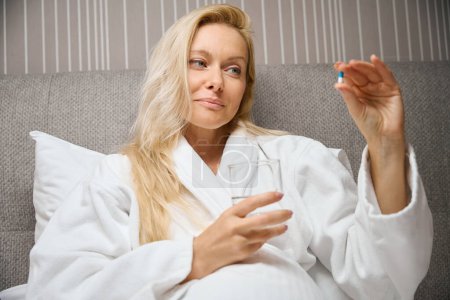 Waist-up portrait of female in bathrobe seated in bed holding glass of water and looking at drug capsule in hand