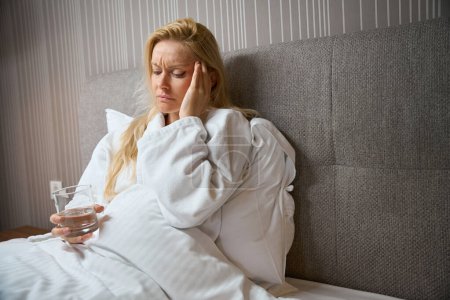 Photo for Despondent female with glass of water seated in bed in bedroom touching her temple with hand - Royalty Free Image