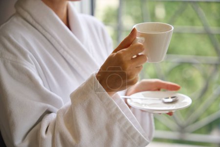 Photo for Cropped photo of lady dressed in bathrobe holding ceramic saucer and cup in her hands - Royalty Free Image