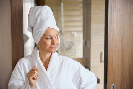 Photo for Portrait of pleased dreamy lady in bathrobe and towel wrapped around head holding toothbrush in hand and looking aside - Royalty Free Image