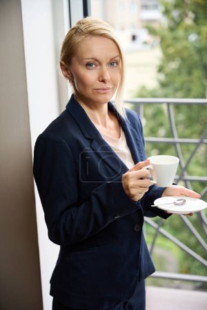 Téléchargez les photos : Élégante dame d'affaires avec soucoupe en céramique et tasse de boisson dans les mains debout dans la porte du balcon dans la chambre d'hôtel - en image libre de droit