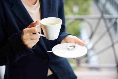 Photo for Cropped photo of elegant businesswoman with ceramic saucer and cup of coffee in hands standing on hotel room balcony - Royalty Free Image