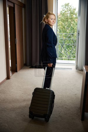 Téléchargez les photos : Portrait grandeur nature d'une femme d'affaires élégante avec des bagages regardant autour dans sa suite à l'arrivée le matin - en image libre de droit