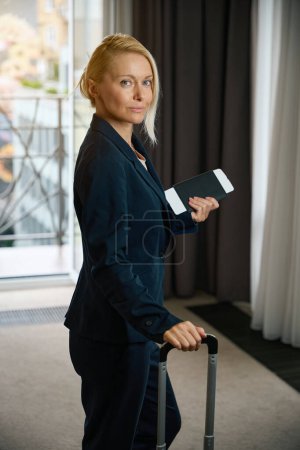 Foto de Elegante mujer de negocios con documentos de viaje y equipaje apoyado en el asa de maleta telescópica en la habitación de hotel - Imagen libre de derechos
