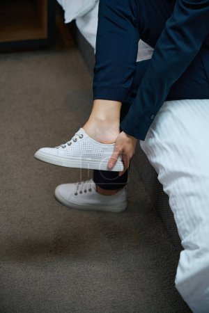 Foto de Foto recortada de la señora en traje de pantalón sentado en la cama en la habitación tirando de zapatillas de cuero blanco - Imagen libre de derechos