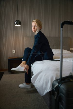 Foto de Mujer empresaria agotada en traje de pantalón quitándose las zapatillas de cuero mientras está sentada en la cama en la suite a su llegada - Imagen libre de derechos