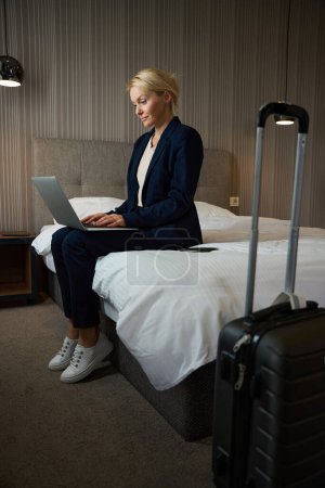 Foto de Mujer de negocios enfocada en traje de pantalón sentada en la cama en la habitación de hotel escribiendo en su computadora portátil - Imagen libre de derechos