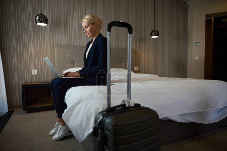 Téléchargez les photos : Femme entrepreneure concentrée vêtue d'une combinaison assise sur le lit en suite tout en tapant sur un ordinateur portable - en image libre de droit