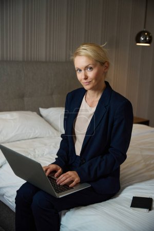 Photo for Business lady in stylish pantsuit sitting on bed in suite while typing on portable computer - Royalty Free Image