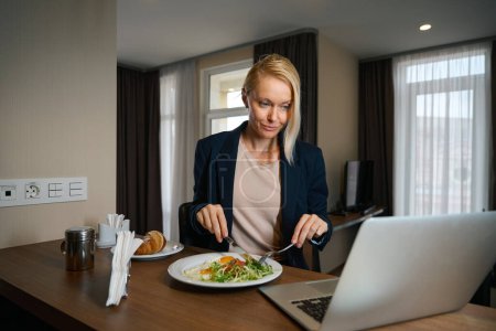 Foto de Mujer emprendedora seria sentada en la mesa en la habitación de hotel cortando comida en el plato mientras mira la pantalla del portátil - Imagen libre de derechos