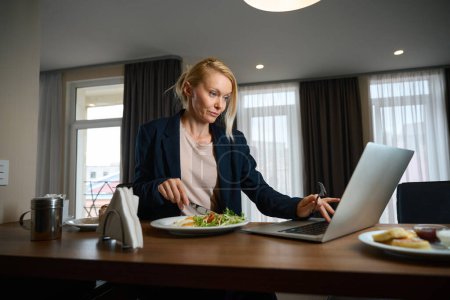 Téléchargez les photos : Femme d'affaires concentrée assise à la table dans la suite couper la nourriture sur la plaque tout en travaillant sur un ordinateur portable - en image libre de droit