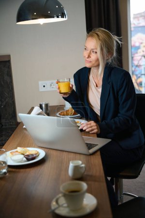 Photo for Dreamy businesswoman with fork and glass of orange juice in hands sitting at table in front of laptop during lunchtime - Royalty Free Image