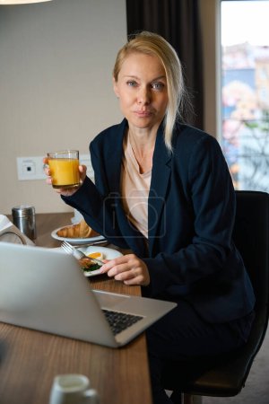 Téléchargez les photos : Femme d'affaires avec fourchette et verre de jus d'orange dans les mains assis à la table devant l'ordinateur portable - en image libre de droit