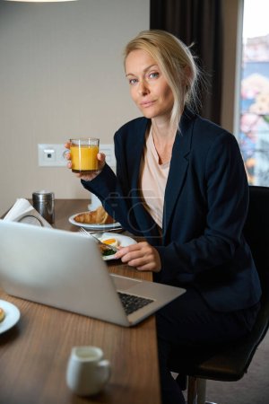 Téléchargez les photos : Femme entrepreneur avec un verre de jus d'orange et fourchette dans les mains assis à la table devant un ordinateur portable au moment des repas - en image libre de droit