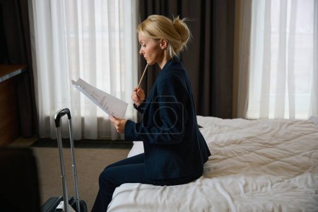 Photo for Side view of focused female entrepreneur with pencil and documents in hands sitting on bed in her suite - Royalty Free Image