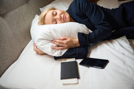 Foto de Mujer de negocios agotada en chaqueta de traje dormitando sobre una almohada suave en una cómoda cama en suite - Imagen libre de derechos