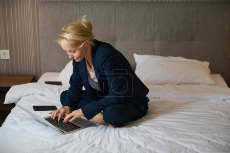 Photo for Focused business lady seated cross-legged on bed in suite typing on her portable computer - Royalty Free Image