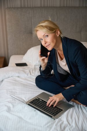 Téléchargez les photos : Femme d'affaires calme assis sur le lit dans la suite tout en tapant sur le clavier de l'ordinateur portable pendant la conversation téléphonique - en image libre de droit
