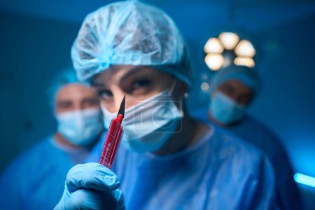 Photo for Young woman doctor wearing uniform and gloves showing scalpel in surgery room interior - Royalty Free Image