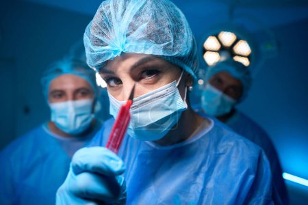 Photo for Professional woman surgeon in hospital operating theater wearing sterile uniform and medical mask showing scalpel - Royalty Free Image