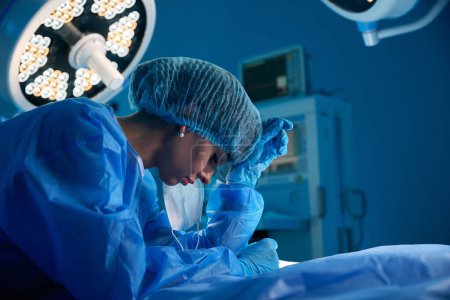 Téléchargez les photos : Profil de la chirurgienne debout dans la salle d'opération après l'opération épuisée et fatiguée - en image libre de droit