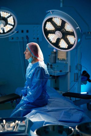 Photo for Exhausted woman surgeon sitting on operating table in operating room after surgery - Royalty Free Image