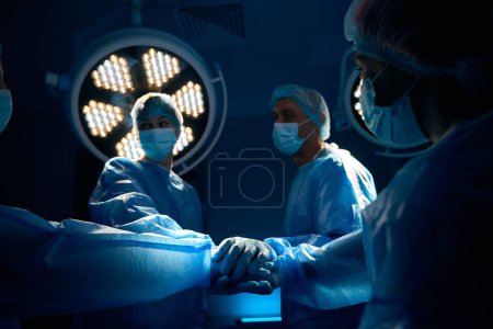 Téléchargez les photos : Médecins chirurgiens équipe tenant la main avant l'opération dans la salle d'opération sombre - en image libre de droit