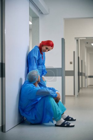 Téléchargez les photos : Femme médecin soutient son collègue après une opération difficile assis dans le couloir de l'hôpital - en image libre de droit