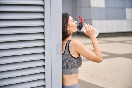Foto de Deportiva hembra cansada después de un duro entrenamiento, parada afuera, sosteniendo una botella de agua y bebiendo. Buenos días deporte - Imagen libre de derechos