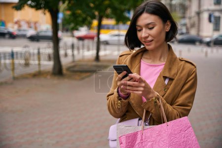 Téléchargez les photos : Femme heureuse avec des sacs d'achat marchant le long de la rue et envoyant des SMS sur smartphone - en image libre de droit