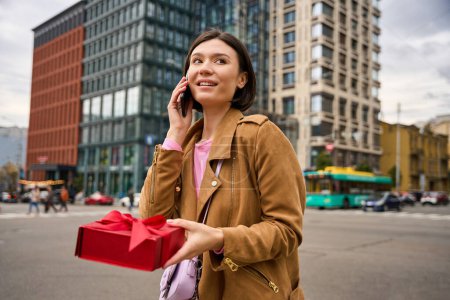 Téléchargez les photos : Femme souriante parlant au téléphone et tenant boîte cadeau rouge avec ruban arc tout en se tenant debout sur la rue de la ville - en image libre de droit