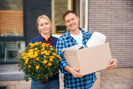 Téléchargez les photos : Tour de taille image d'un homme joyeux portant une boîte en carton et une femme souriante tenant des fleurs lorsqu'elle se tient près du bâtiment pendant le déménagement de la maison - en image libre de droit