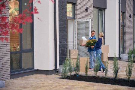 Photo for Contented man carrying cardboard box and happy lady holding flowerpot with flowers while couple walking towards new house - Royalty Free Image