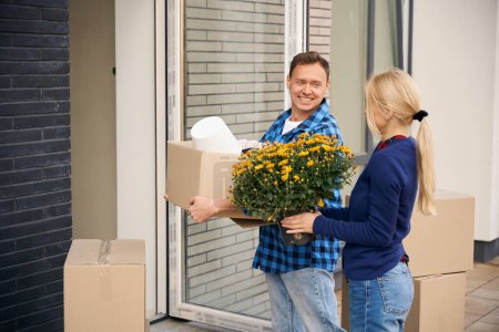 Téléchargez les photos : Couple heureux debout près de l'entrée de la maison tout en se regardant et en portant leurs affaires dans les mains - en image libre de droit