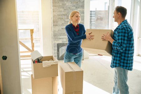Laughing couple standing in new home when man giving cardboard box to woman and she taking it
