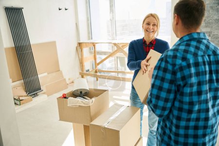 Cheerful lady giving box to man who standing near her and helping to unpack boxes after house move