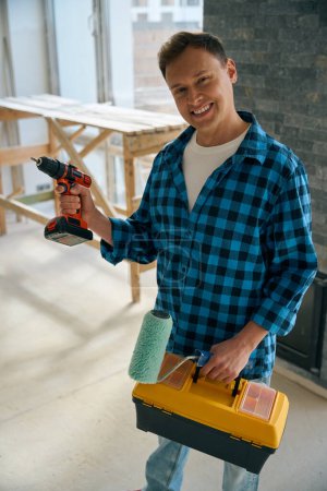 Foto de Hombre de pie en la habitación mientras sostiene el kit de herramientas y el rodillo de pintura en la mano izquierda, mientras que el destornillador eléctrico en el derecho - Imagen libre de derechos