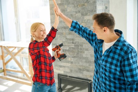 Waist up picture of cheerful lady and man standing in room and giving high five to each other