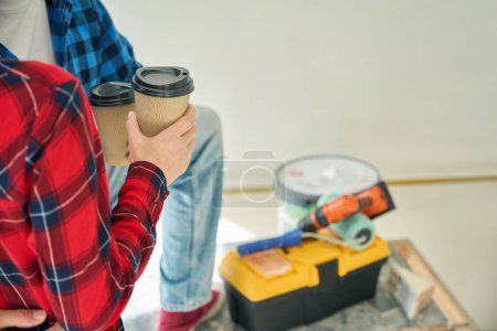 Cropped photo of man and woman hands holding coffee in ripple paper glasses while construction tools lying in back ground