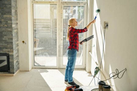 Photo for Side view photo of smiling workwoman painting wall with paint roller while standing in empty room of new house - Royalty Free Image