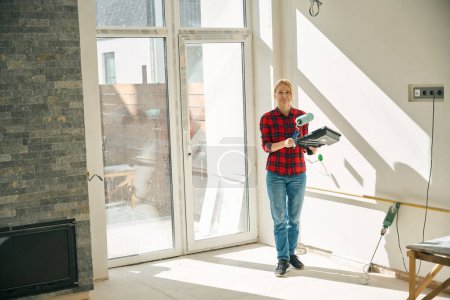 Foto de Foto de larga duración de la mujer alegre de pie en una habitación incompleta mientras sostiene la bandeja de pintura y el rodillo en las manos - Imagen libre de derechos