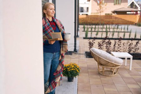 Téléchargez les photos : Femme satisfaite debout à l'extérieur de la maison tout en tenant le livre et la tasse dans les mains et regardant vers l'avenir - en image libre de droit