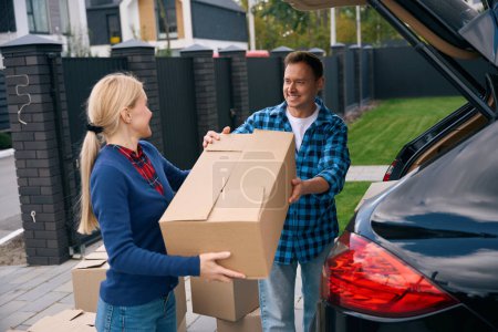 Photo for Smiling man unloading cardboard box out of car trunk and giving it to happy woman standing near him - Royalty Free Image