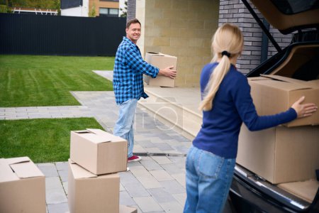 Photo for Lady standing and unloading cardboard box out of car trunk while joyful man carrying another box towards house - Royalty Free Image