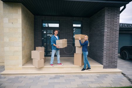Photo for Full length picture of man and lady standing on porch of new house while arranging cardboard boxes - Royalty Free Image