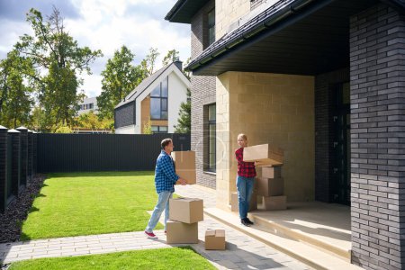Photo for Full length picture of contented couple walking towards house entrance while holding boxes in hands - Royalty Free Image