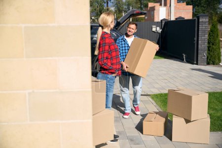 Foto de Señora de pie cerca de la casa cuando el hombre alegre llevando cajas de cartón y organizándolos cerca de la puerta - Imagen libre de derechos